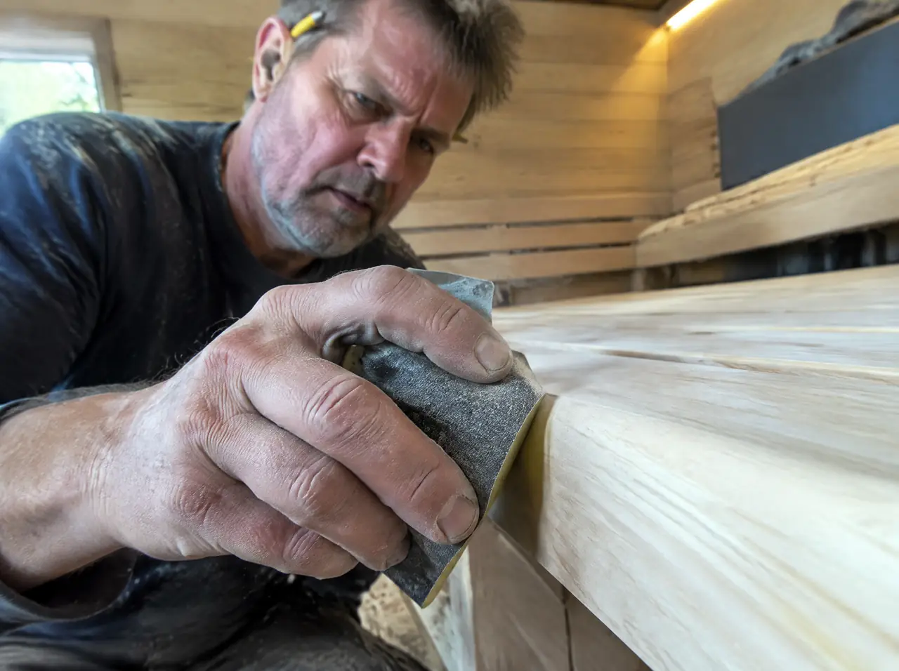 Weathered hands sanding the rounded edge of a pale ash sauna bench in a workshop