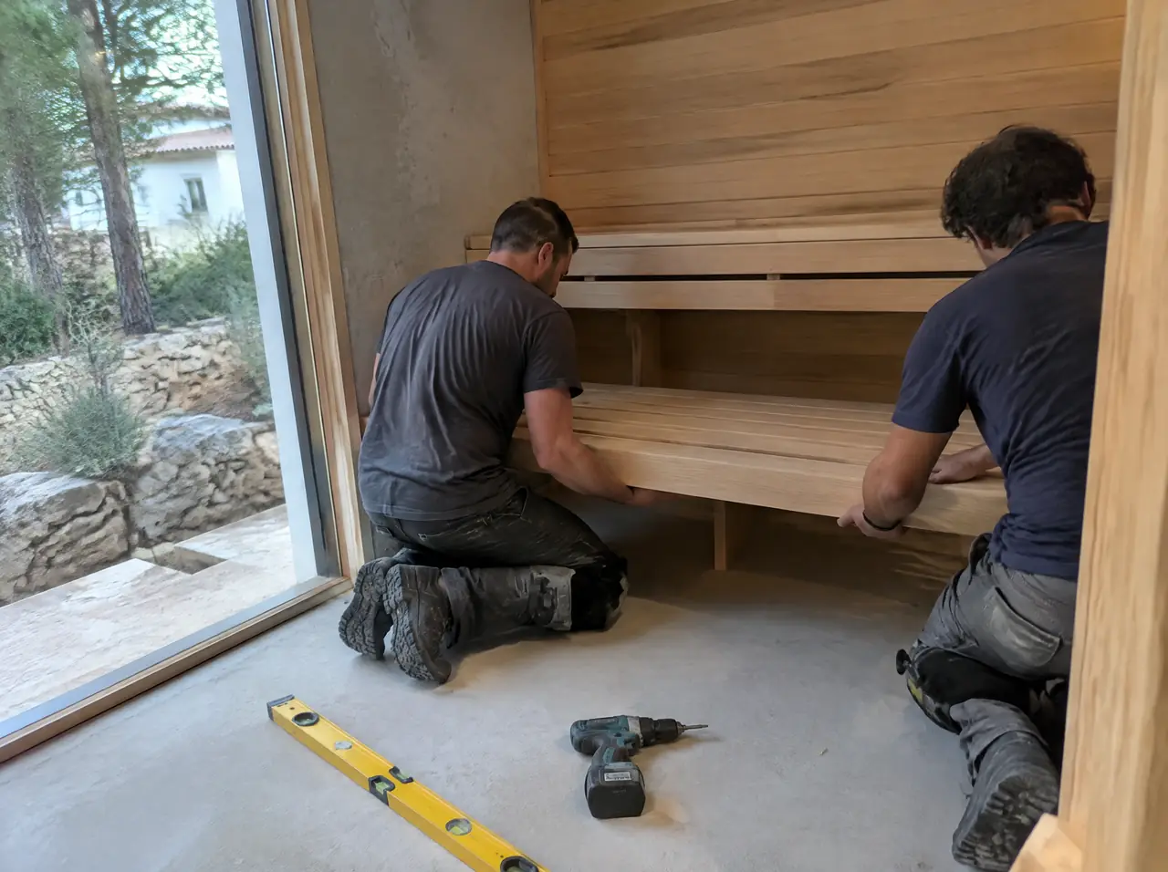 Two installers in knee pads fitting a slatted ash bench inside a half-finished sauna cabin in a Portuguese villa