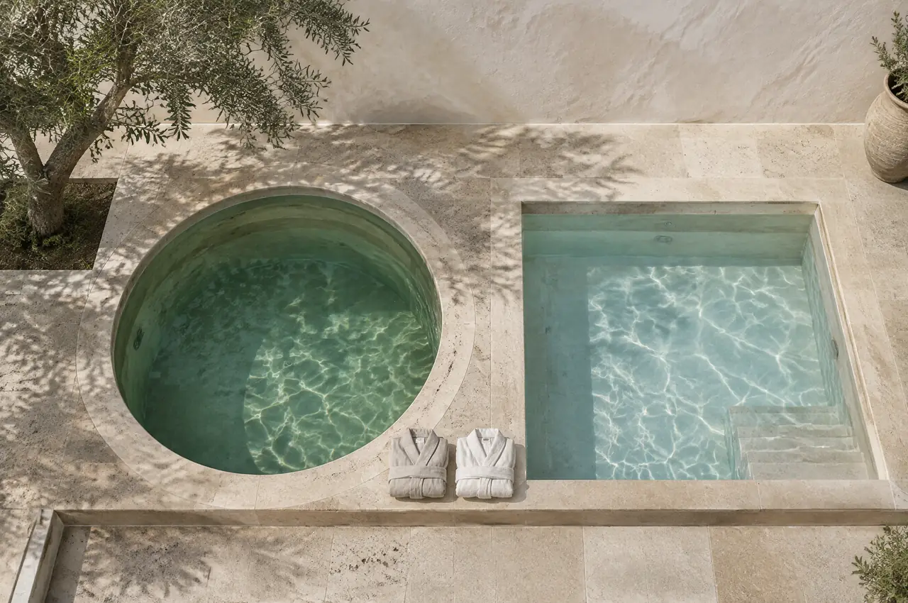 Overhead view of a round warm pool and a square cold pool side by side on a travertine terrace with two folded robes