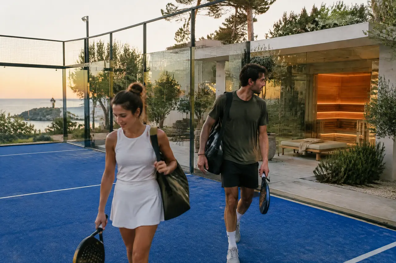 Padel court at dusk with blue turf and glass walls, a glass-fronted recovery pavilion with a sauna glowing behind and two players walking off court