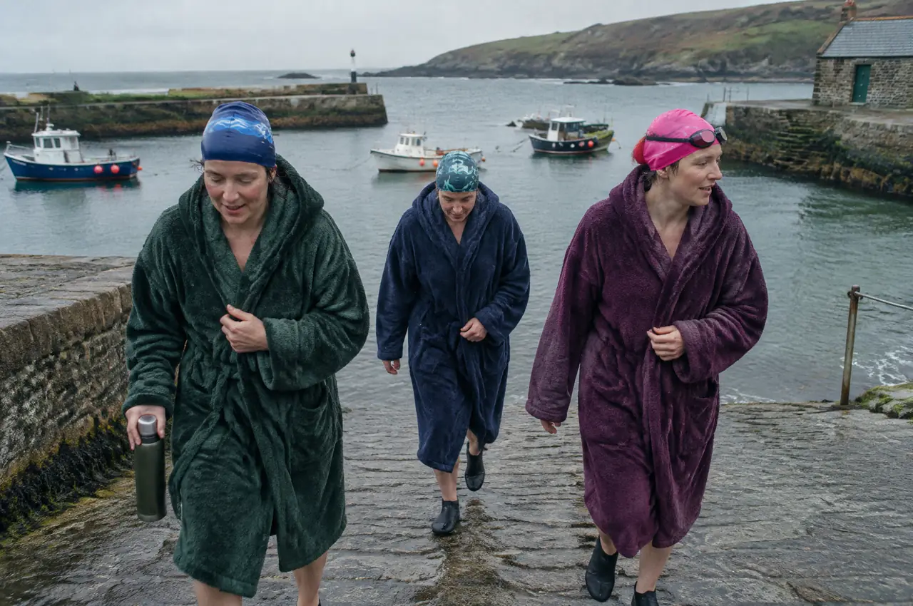 Three Irish sea swimmers in hooded towelling robes and swimming caps walking up a stone slipway from a grey-green harbour, one holding a flask
